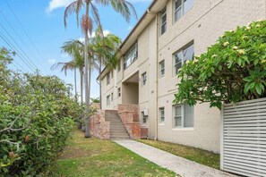 A multi-story residential building with a brick facade, a concrete pathway leading to the entrance, and a small garden area with palm trees and shrubs. - The Little Lawrence Sunny Village Nook by the Sea (Freshwater)