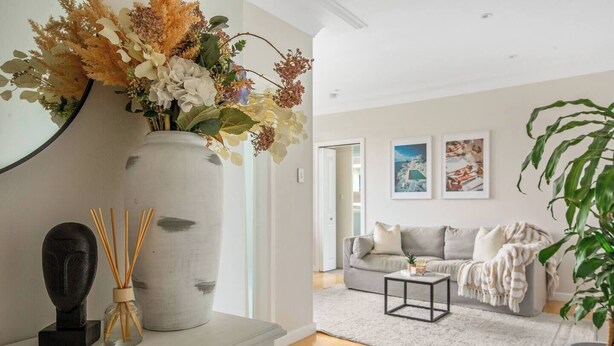 A modern living room with a large vase of flowers, a black sculpture, and a small table with a candle. There is a grey sofa with a throw blanket and a potted plant. - The Little Lawrence Sunny Village Nook by the Sea (Freshwater)