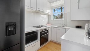 A modern kitchen with white cabinetry, a built-in water dispenser, a gas stove, an oven, a microwave, and a window with a view of palm trees. - The Little Lawrence Sunny Village Nook by the Sea (Freshwater)