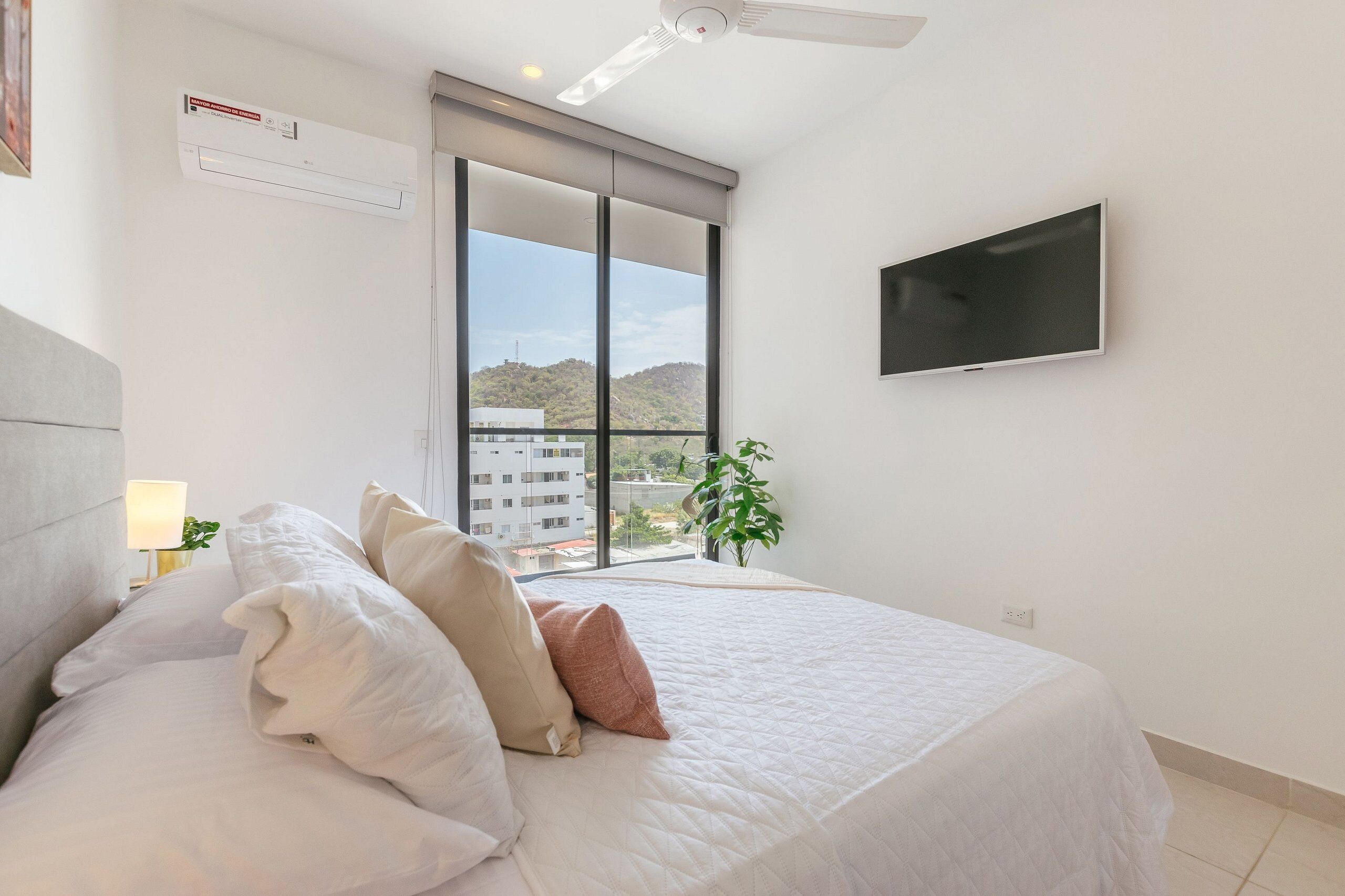 Een slaapkamer met een bed, een flatscreen televisie, een airconditioningapparaat, een raam met uitzicht en een plant.
