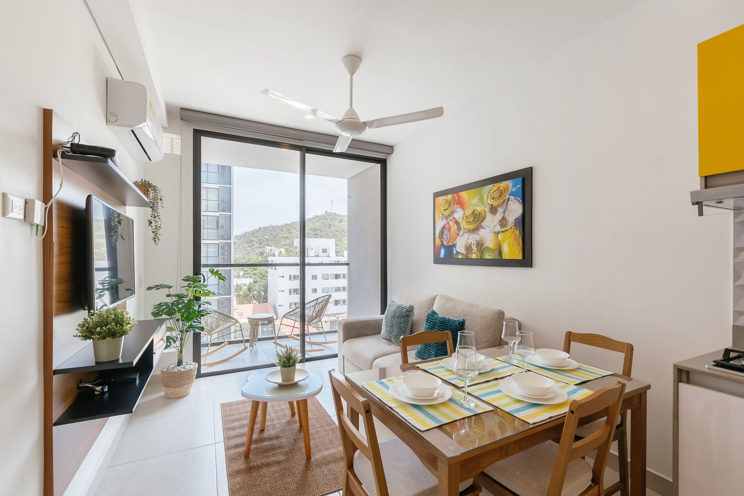 A modern living room with a dining area, large windows, a ceiling fan, and a painting on the wall.
