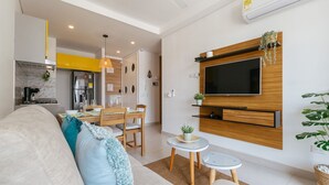 Un salon moderne avec un canapé, un téléviseur fixé au mur, un coin repas et une cuisine en arrière-plan.
