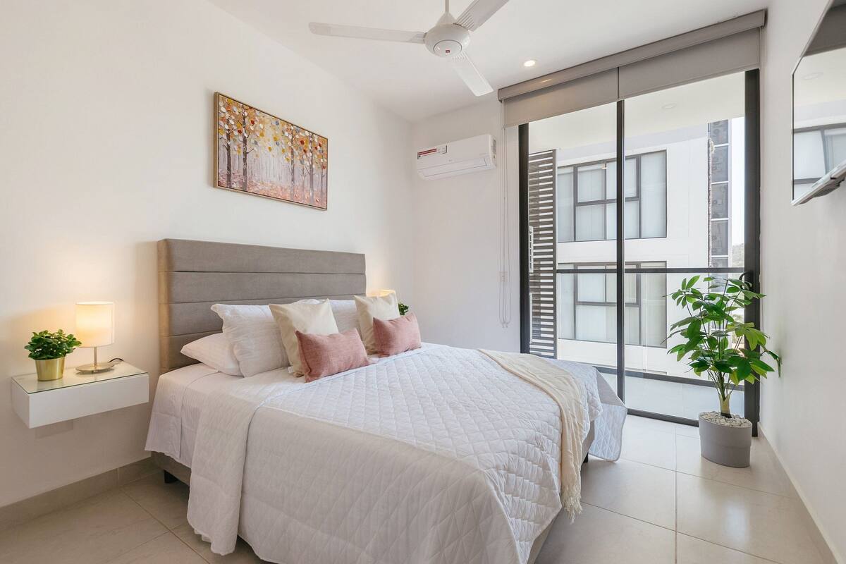 Une chambre à coucher comprenant un lit, des tables de chevet, un ventilateur de plafond, une fenêtre avec des stores et une plante en pot.