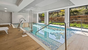 Una piscina interior moderna con paredes de vidrio transparente, suelo de madera y vistas a un paisaje rocoso.