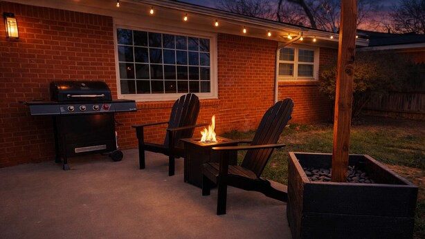 Ein Garten mit einer Ziegelmauer, einem Grill, zwei Adirondack-Stühlen, einer Feuerstelle und Lichterketten.