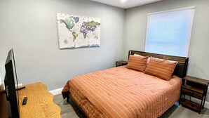 Ein Schlafzimmer mit Bett, Schreibtisch mit Computer, einer Weltkarte an der Wand und einem Fenster mit Jalousien.
