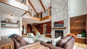 A spacious living area with a stone fireplace, wooden beams, and a staircase leading to an upper level. - Newly Constructed Lake Access Home w/ Private Pool (Swanton)