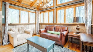 A cozy room with a wooden ceiling, large windows, a brown leather sofa, a beige armchair, and a wooden coffee table. - New! Cute Hideaway w/ Hot Tub & Amenities (Oakland)