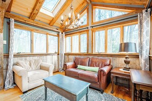 Ein gemütliches Zimmer mit Holzbalkendecke, großen Fenstern, einem braunen Ledersofa, einem beigefarbenen Sessel und einem Holztisch.