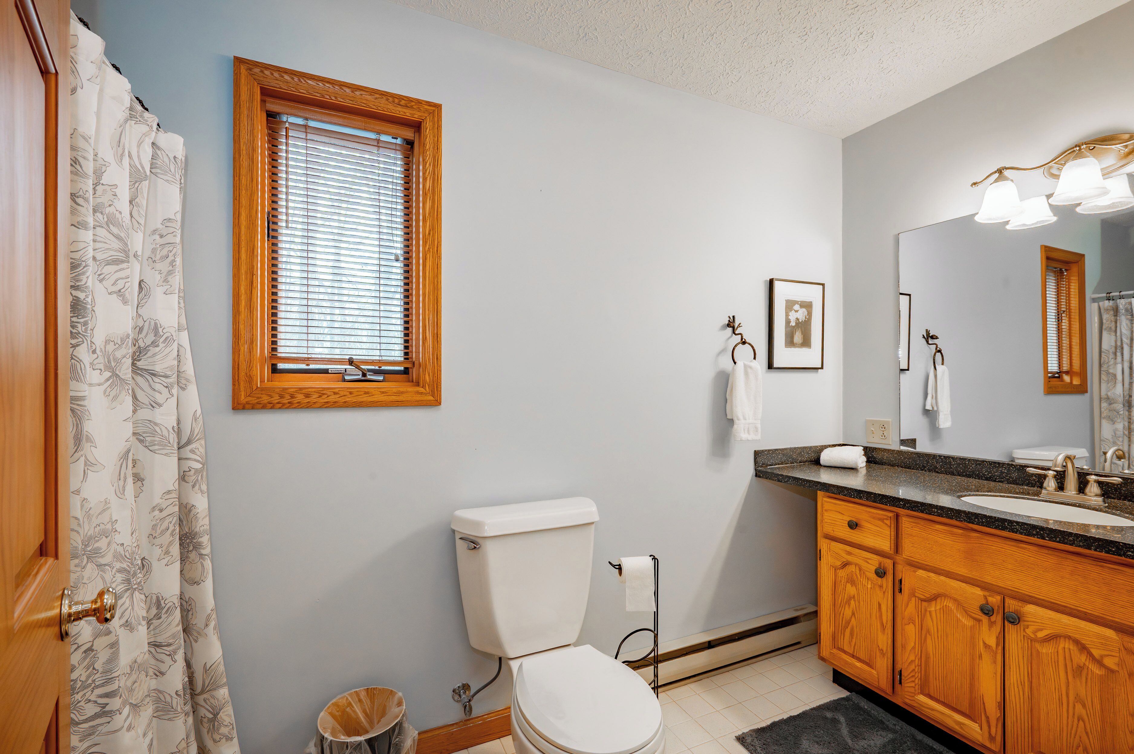 A bathroom with a toilet, sink, wooden cabinets, a window with blinds, and a shower curtain.