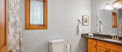 A bathroom with a toilet, sink, wooden cabinets, a window with blinds, and a shower curtain.