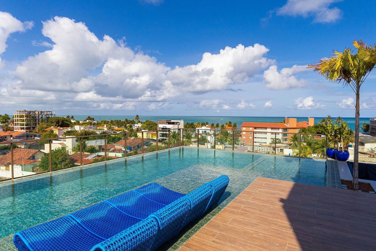 A clear blue pool with a blue lounge chair, surrounded by a wooden deck and palm trees, overlooking a coastal town.
