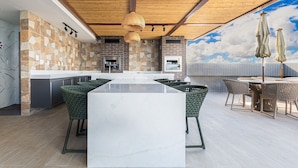A modern kitchen with a large island, stone backsplash, and outdoor seating area with wicker chairs and umbrellas. - Makani Beach #07 - Flat com Piscina by Carpediem (Tamandaré)