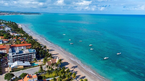 An aerial view of a coastal area with a beach, several boats, and a hotel with multiple floors and balconies. - Makani Beach #07 - Flat com Piscina by Carpediem (Tamandaré)