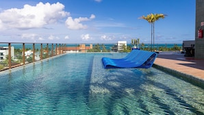 A clear blue pool with a blue lounge chair, surrounded by a metal railing and palm trees, with a view of the ocean and buildings in the background. - Makani Beach #07 - Flat com Piscina by Carpediem (Tamandaré)