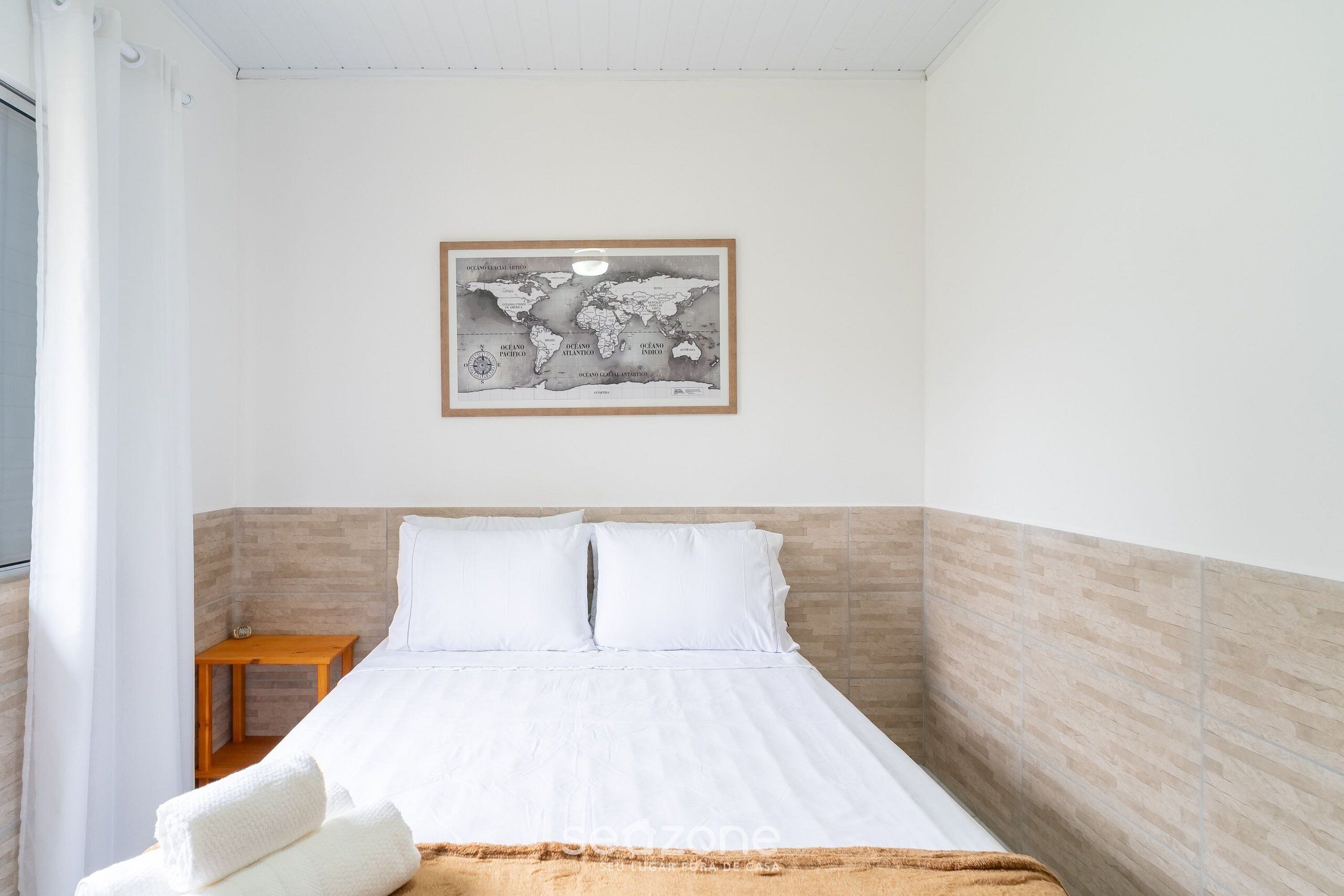 A neatly made bed with white linens, a small wooden nightstand, a world map artwork on the wall, and a light-colored tiled wall.