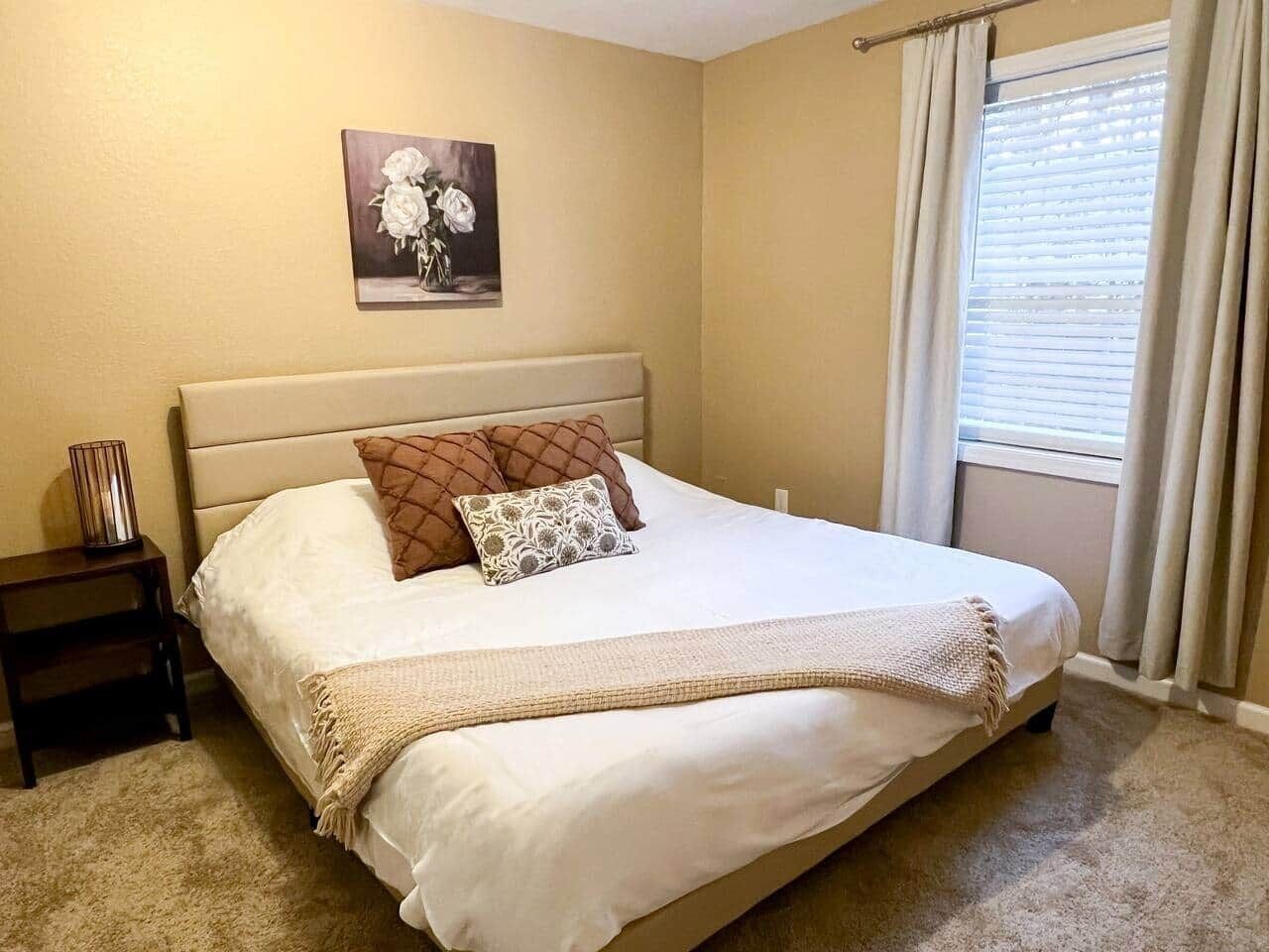 Une chambre à coucher avec un lit, une table de chevet, un tableau au mur et une fenêtre avec des rideaux.
