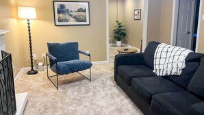 Un salon avec un fauteuil bleu, un canapé noir recouvert d’un plaid, un lampadaire et un tableau représentant un paysage, accroché au mur.
