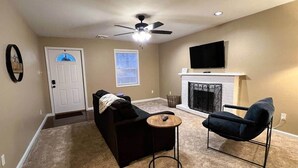 Un salon avec un canapé noir, un fauteuil bleu, une petite table ronde, une cheminée et un ventilateur de plafond.