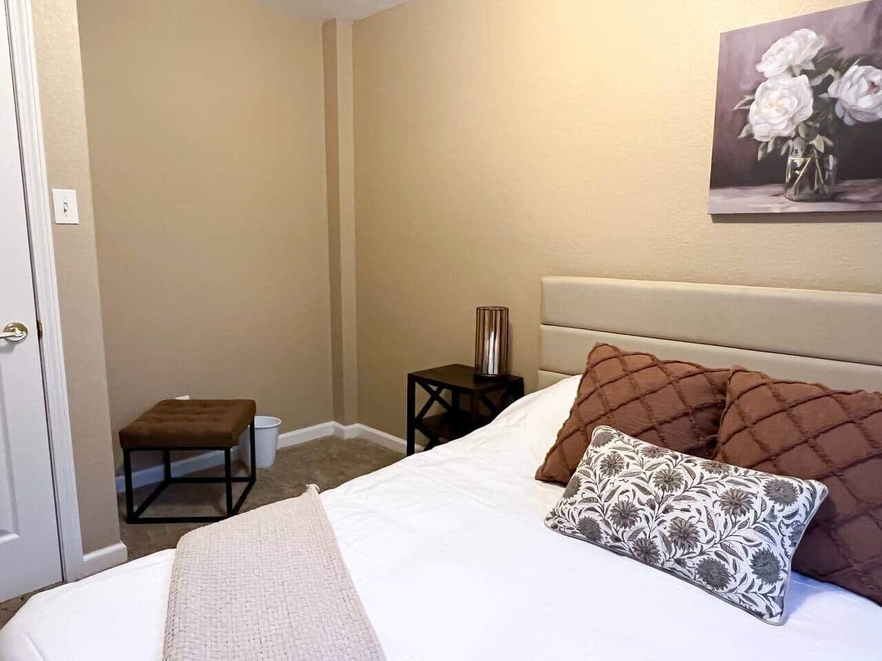 Une chambre à coucher avec un lit, une table de chevet, un petit tabouret et un tableau accroché au mur.