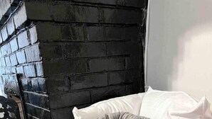 A wicker basket with pillows and a blanket, placed on a wooden floor near a black brick wall.