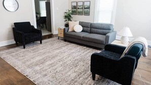 Uma sala de estar com um sofá cinzento, duas poltronas pretas, um tapete estampado, uma pequena mesa lateral com uma luminária e quadros nas paredes.
