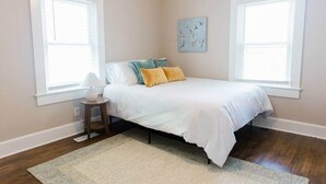 Quarto com cama, duas janelas, mesa de cabeceira e uma obra de arte na parede.