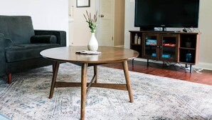 Una sala de estar con una mesa de centro redonda de madera, un sofá gris, un televisor sobre un mueble bajo y un aparador con puertas de cristal.