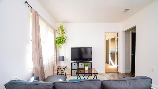 A living room with a sofa, a TV, a small table with a plant, and a lamp. There is a window with curtains and a door leading to another room. - 2 Bedroom Apt in Downtown ATL, Steps from Beltline (Atlanta)