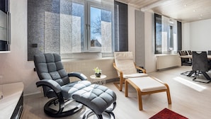 A modern living room with a sofa, armchair, and ottoman, a dining area with a table and chairs, and large windows with patterned curtains. - Apartment 'Messe-und-Ferien-Wohnung' with Wi-Fi (Seelze)