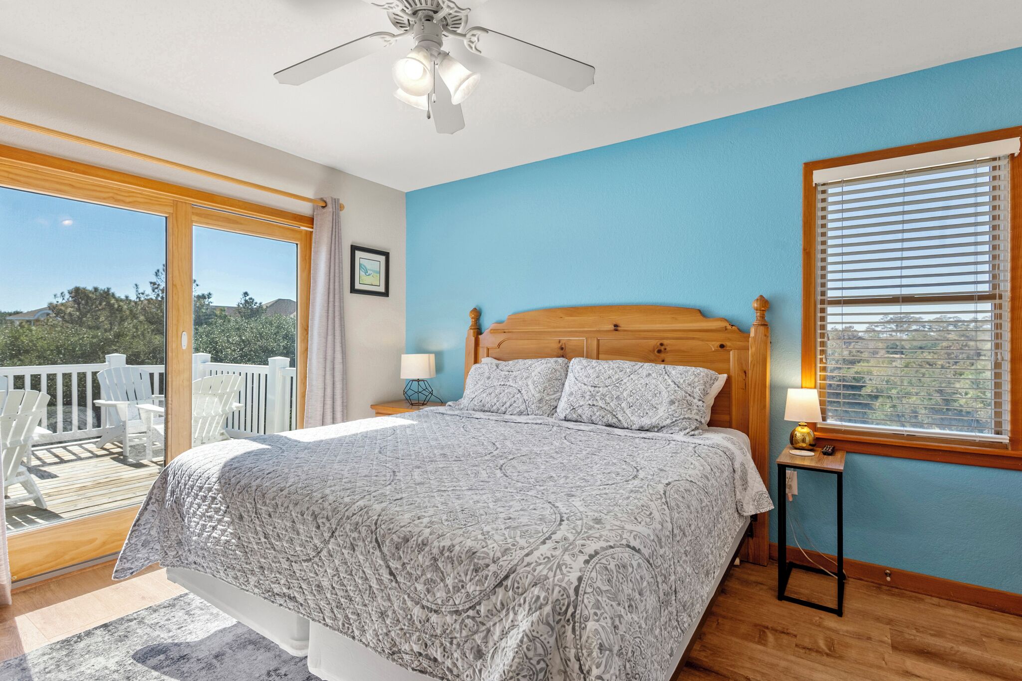 A bedroom with a large bed, a ceiling fan, a window with blinds, and a balcony with white chairs.