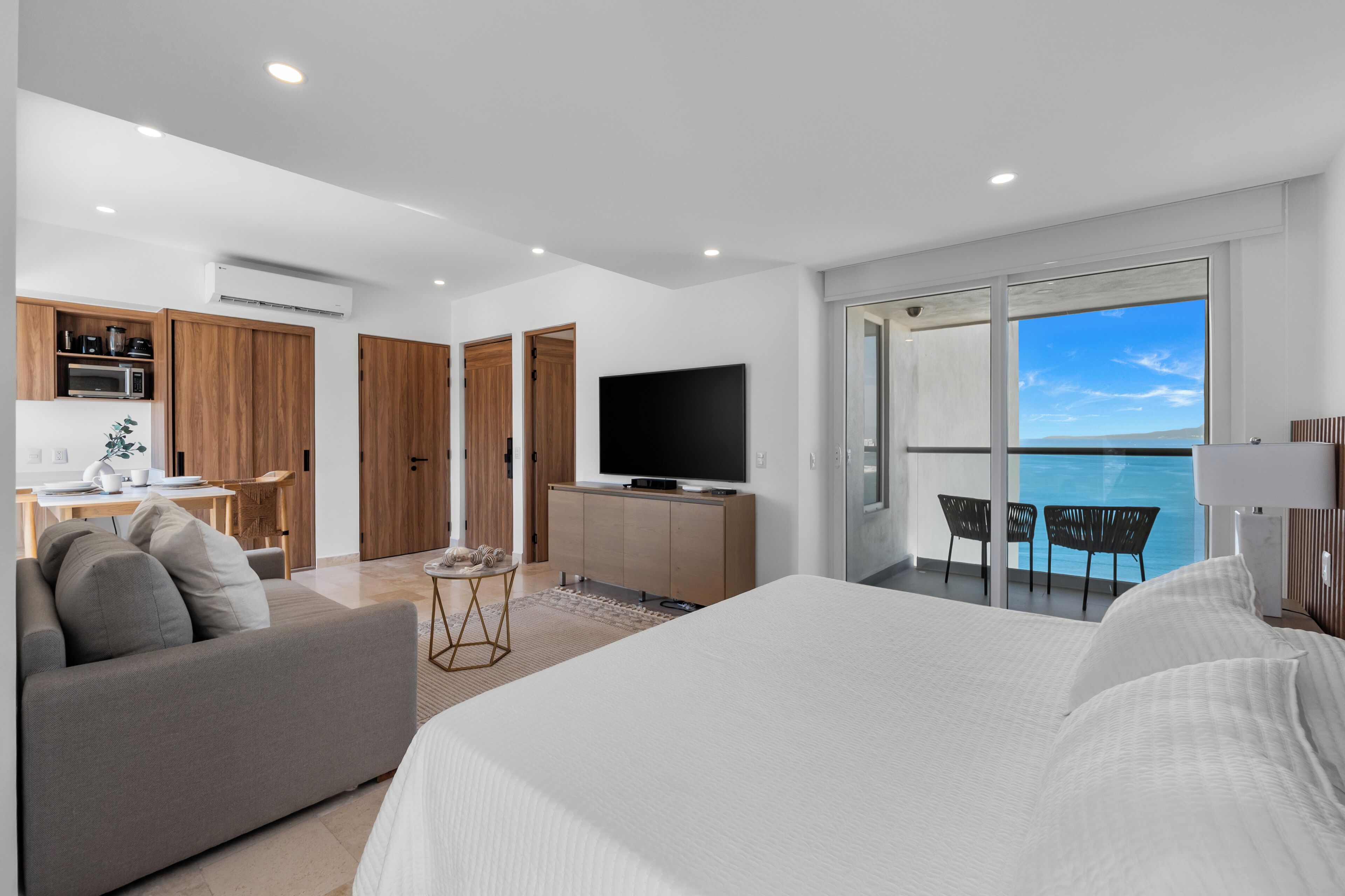 A modern hotel room with a large bed, a TV, a balcony with a view of the sea, and a kitchenette.