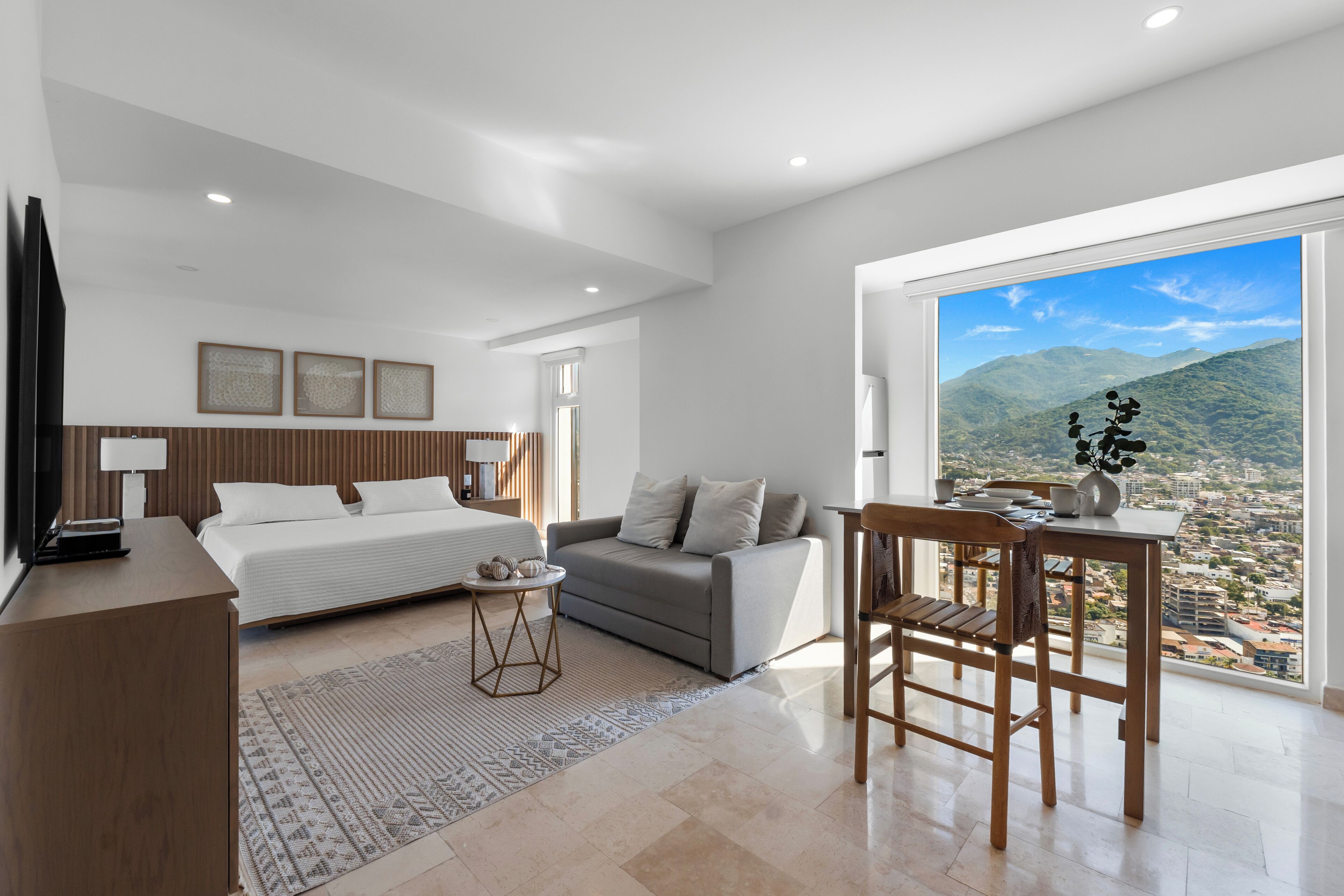 A modern bedroom with a bed, a sofa, a dining table, and a view of a cityscape through large windows.