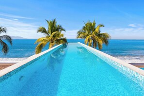 A swimming pool with clear blue water, surrounded by palm trees, leading to a beach and ocean view. - Amazing Apartments - Hotel Zone (Puerto Vallarta)