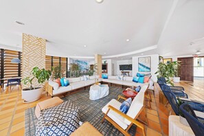 A spacious living area with a large sofa set, a coffee table, and several chairs. There are potted plants and a wall with a geometric pattern.