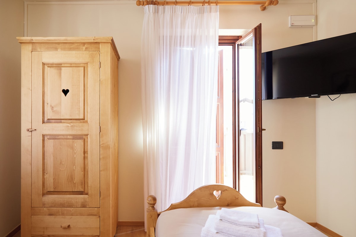 Un dormitorio con un armario de madera, una cama con ropa blanca, un televisor montado en la pared y un ventanal con cortinas traslúcidas.