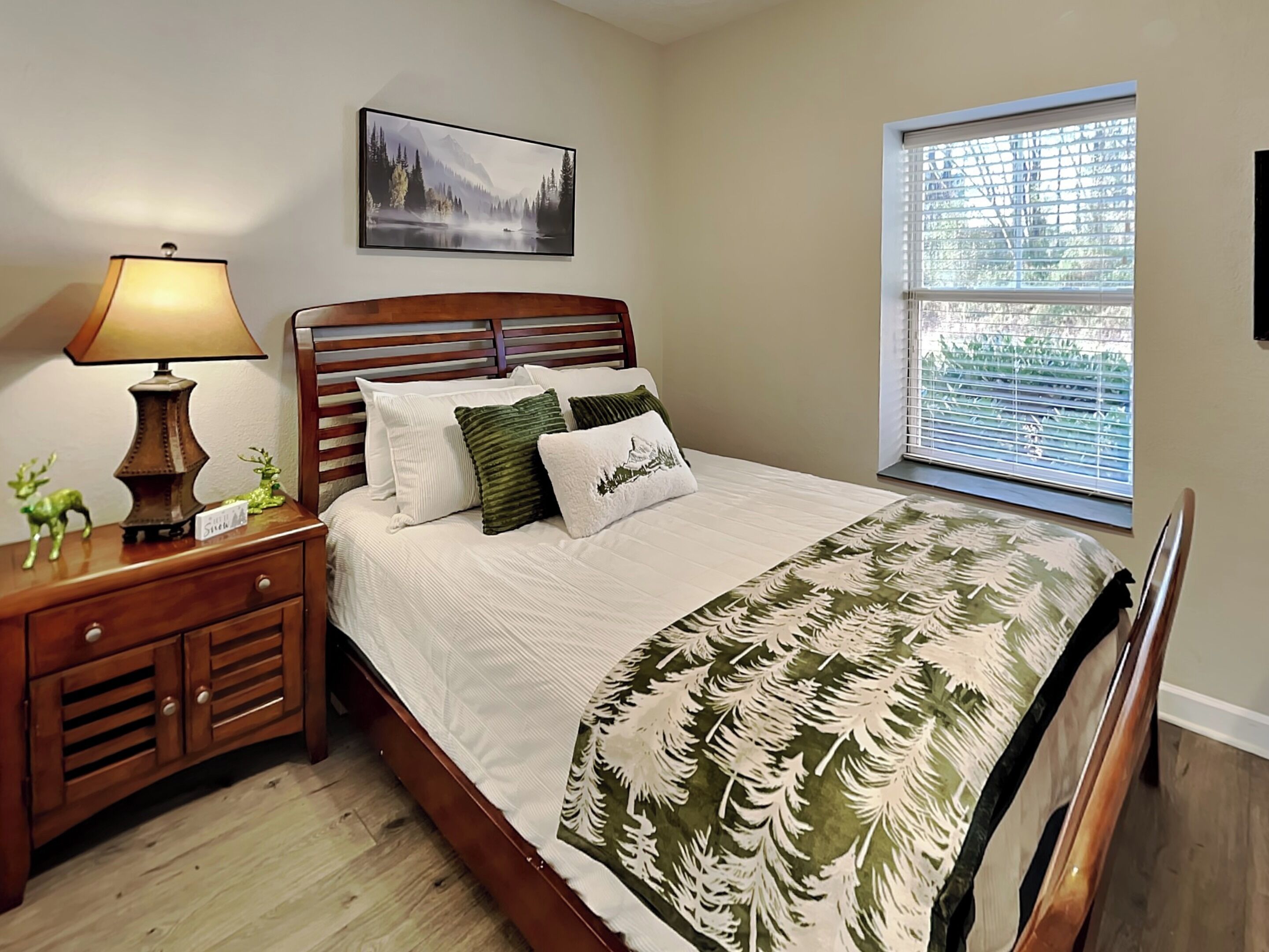 A bedroom with a wooden bed, a nightstand with a lamp, a window with blinds, and a framed picture on the wall.