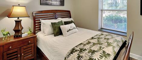 A bedroom with a wooden bed, a nightstand with a lamp, a window with blinds, and a framed picture on the wall.