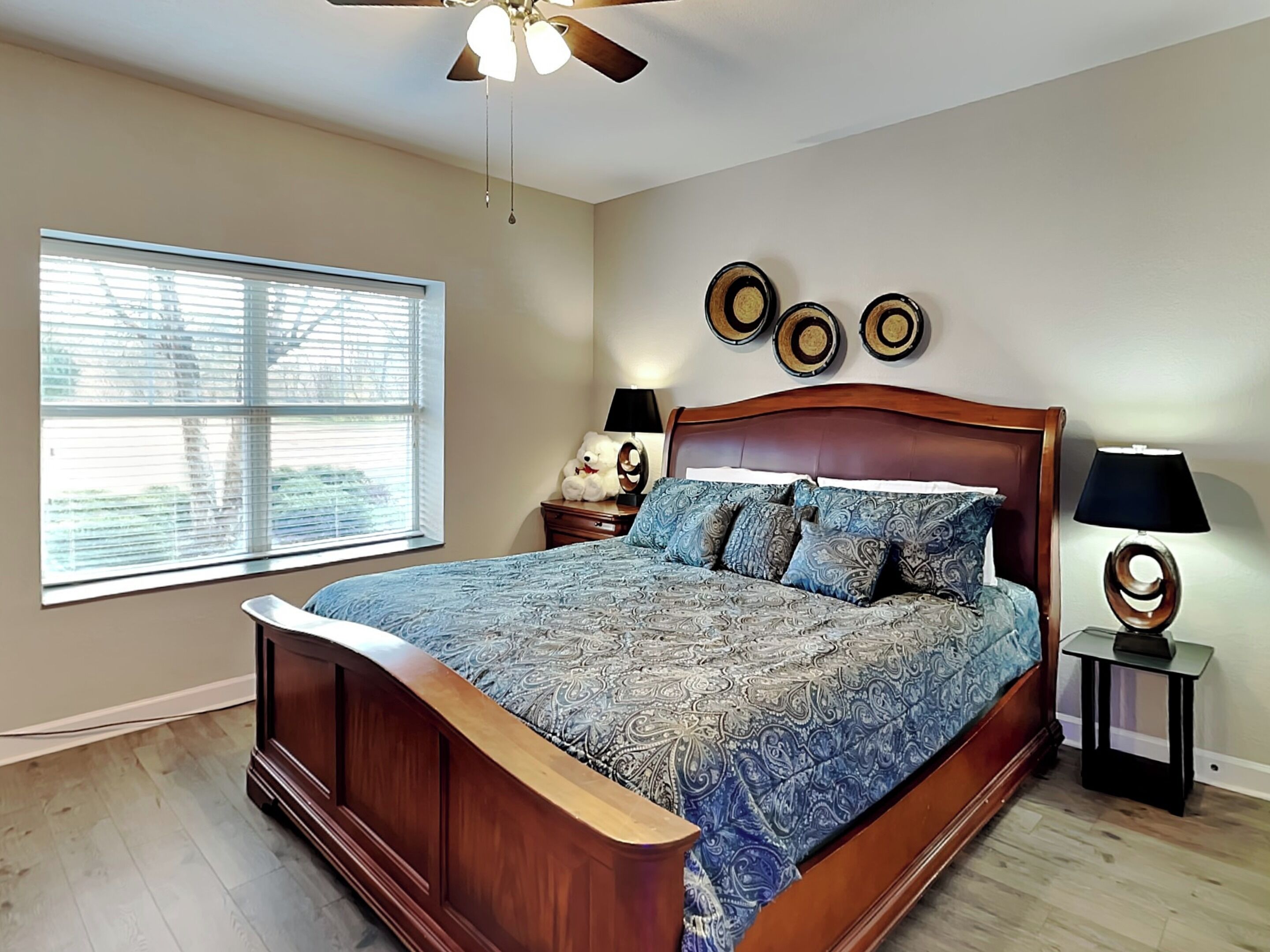 A bedroom with a wooden bed, a large window with blinds, a ceiling fan, and decorative wall plates.