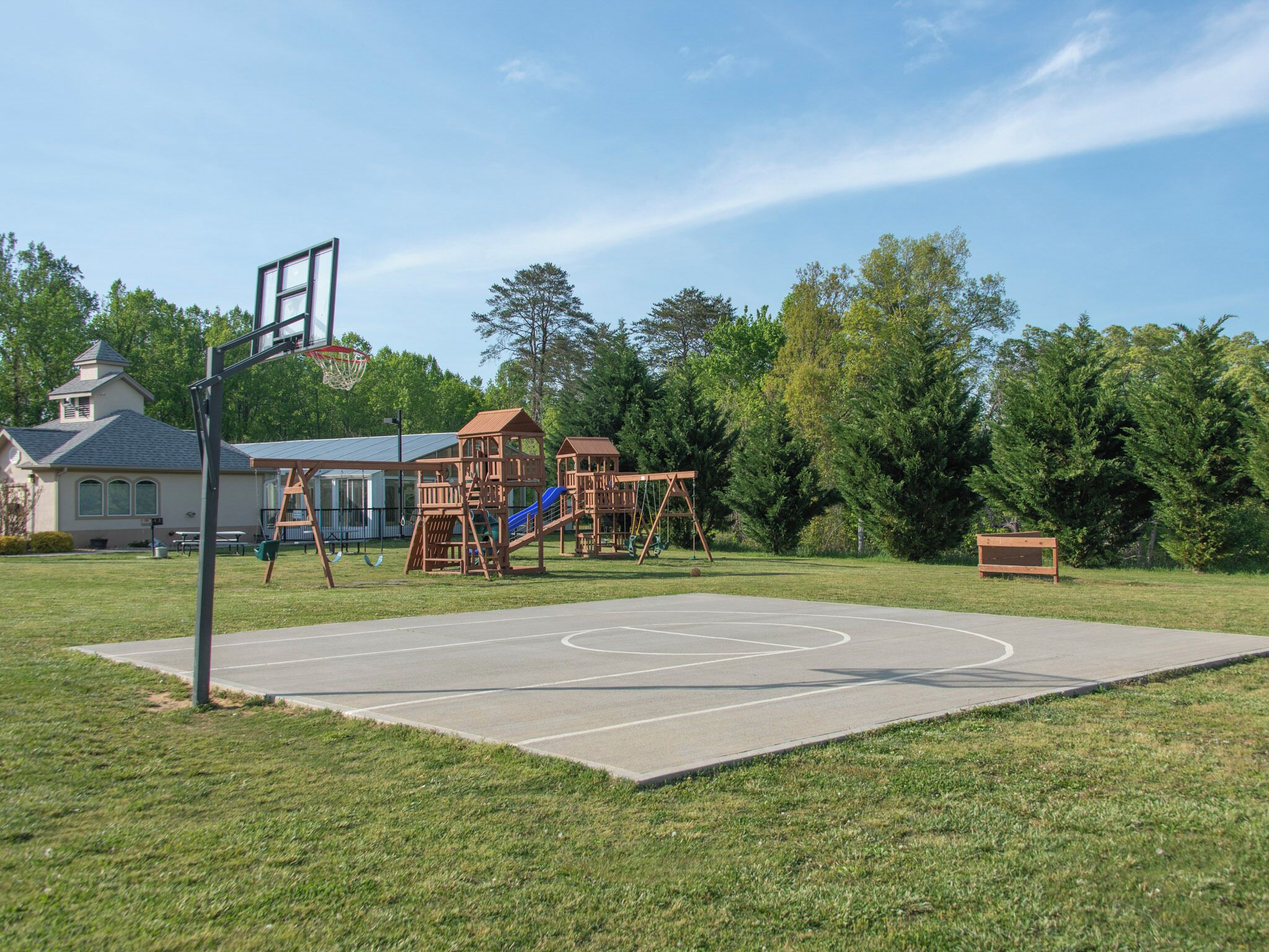 Un parque infantil con canchas de baloncesto, estructuras de juego de madera y una casa al fondo.
