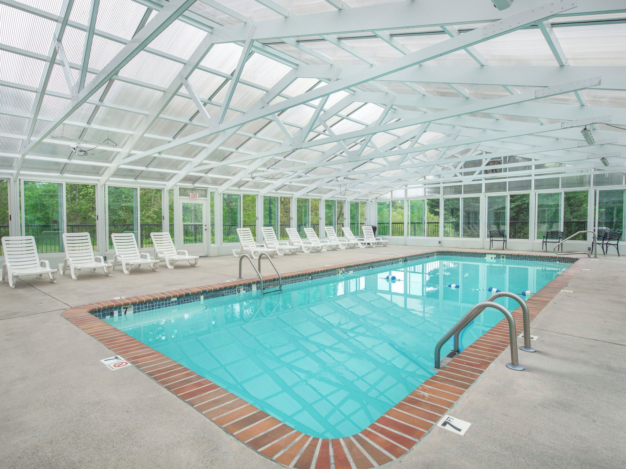 Une piscine intérieure couverte, dotée de parois en verre transparent, de chaises longues blanches et d’un bord de piscine en briques.