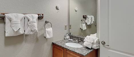 A bathroom with a granite countertop sink, wooden cabinets, a mirror, and multiple white towels hanging on towel racks.