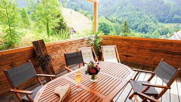 Eine Holzterrasse mit einem runden Tisch und Stühlen, einer Blumenvase und Blick auf ein bewaldetes Tal.