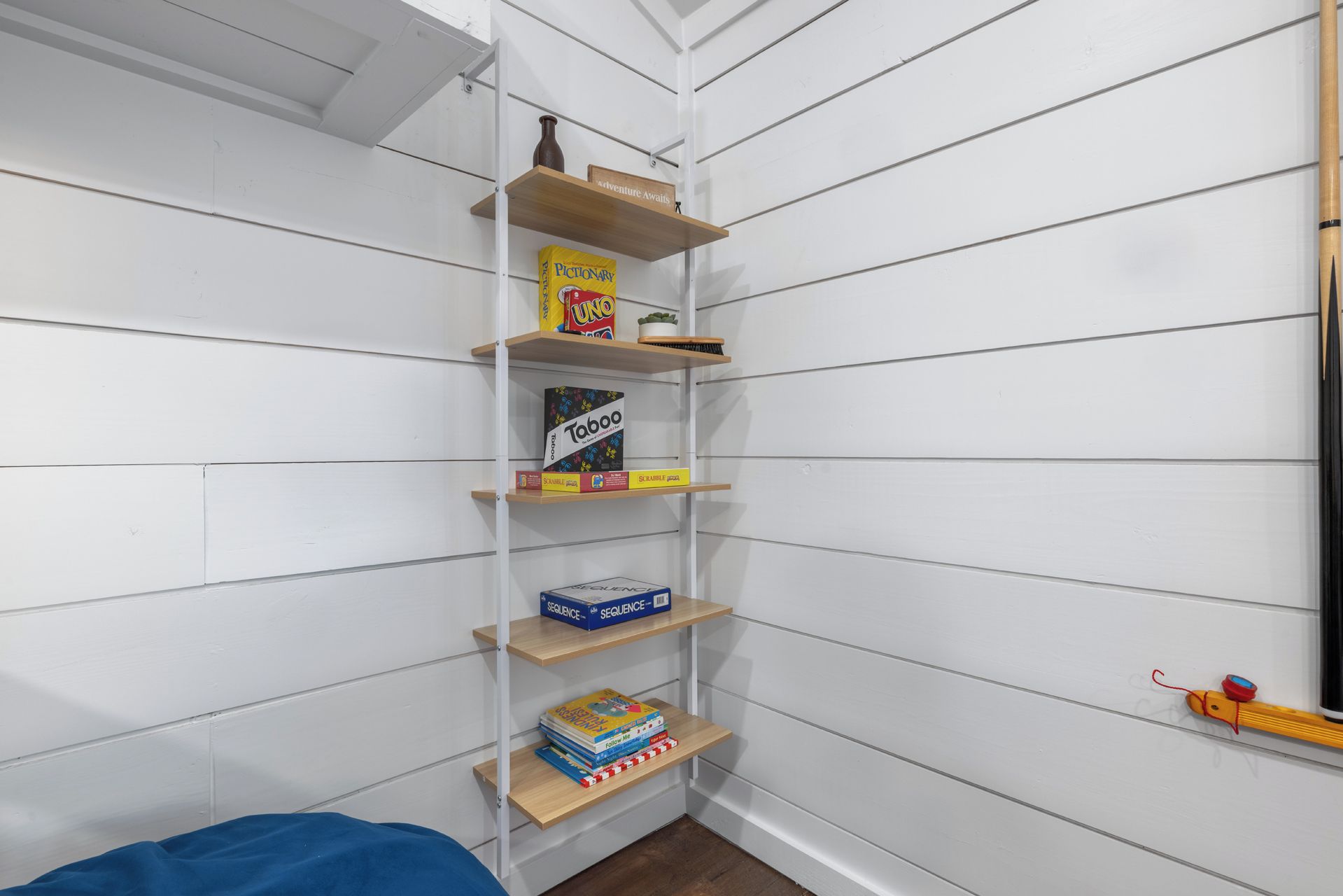 A corner of a room with white paneled walls and a wooden shelf unit. The shelf holds a variety of board games, including 'Uno' and 'Taboo'.