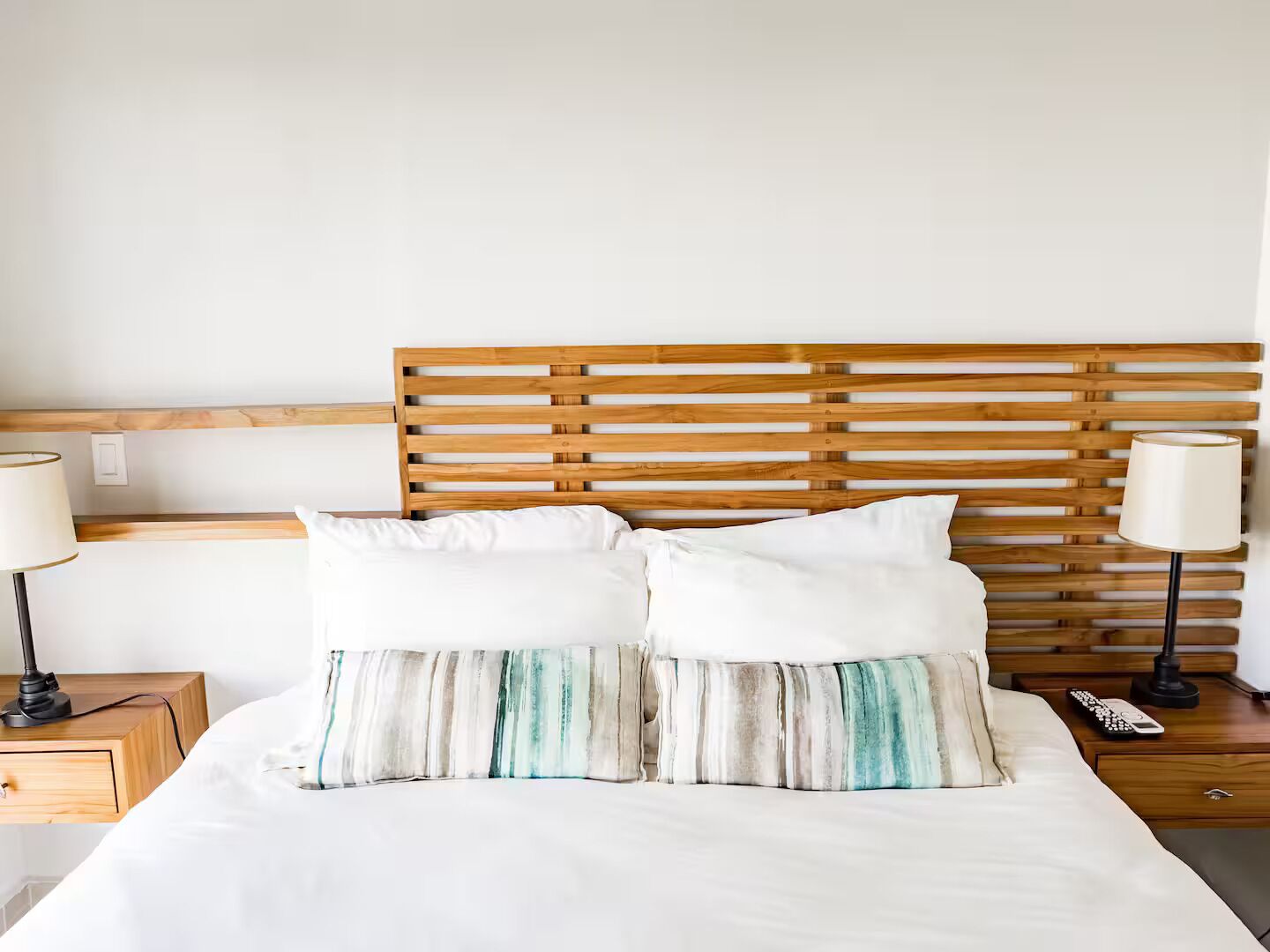 A neatly made bed with white bedding and two pillows, a wooden headboard, and bedside tables with lamps and a remote control.