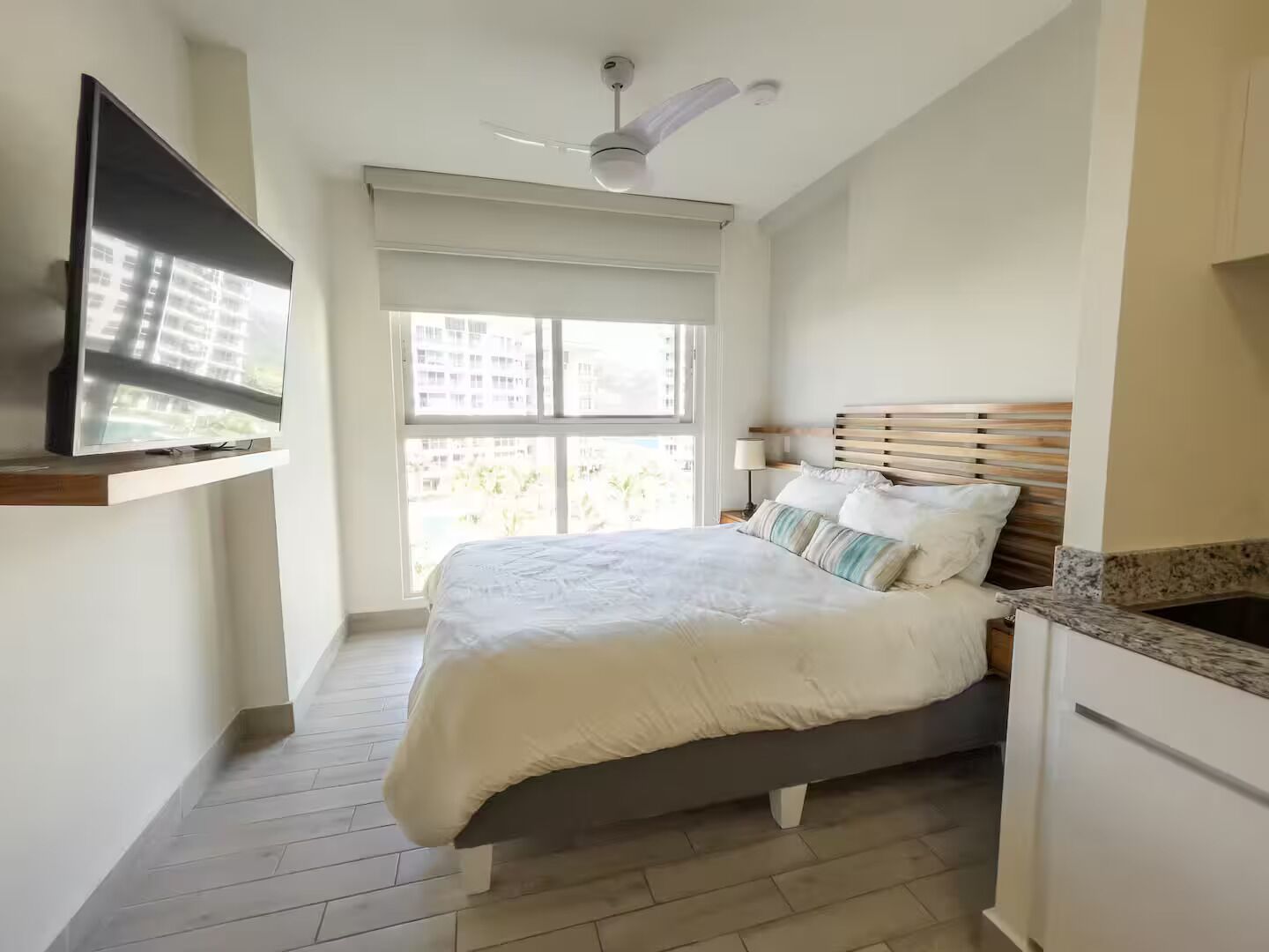 A bedroom with a bed, a TV mounted on the wall, a ceiling fan, and a window with a view of buildings.