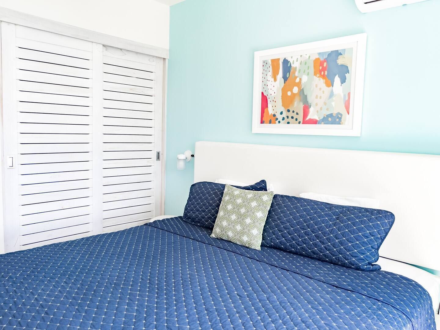 Een slaapkamer met een blauw gebogen beddegoed, een wit hoofdeinde en een kleurrijk abstract schilderij aan de muur.