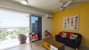 A modern living room with a sofa, coffee table, and chairs, a large window with a view of the ocean, and a ceiling fan. - Sea View Breeze -V3-4C- 2 bed, 2 bath (Punta Chame)