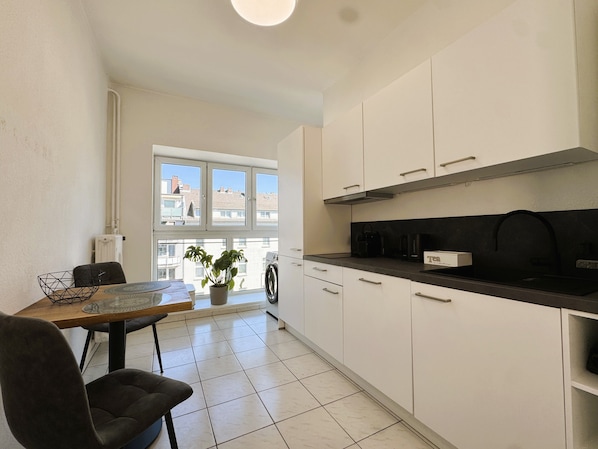 Une cuisine moderne avec des meubles blancs, un plan de travail noir et une fenêtre donnant sur des immeubles.