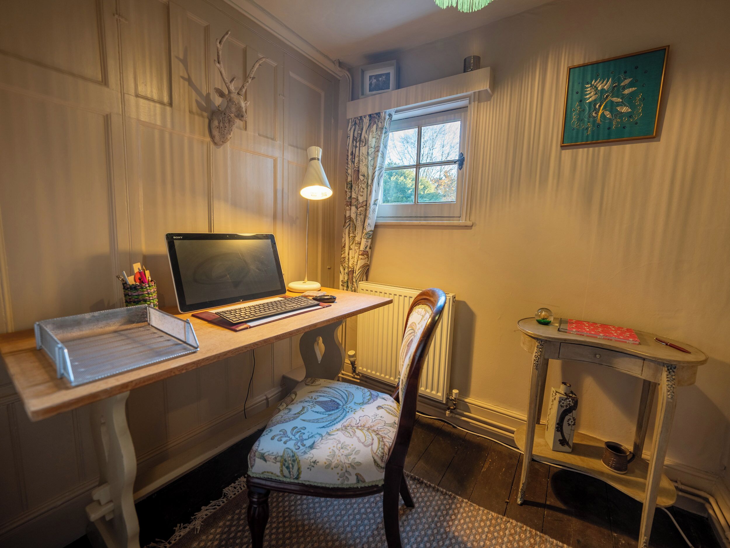A study with a desk, laptop, chair, radiator, and a small table with a lamp.